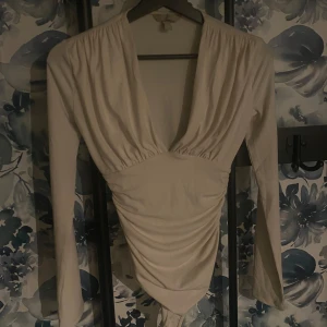 Beige långärmad topp med v-ringning - Säljer en stilren beige topp med lång ärm och v-ringad design. Toppen har en åtsittande passform och är perfekt för både vardag och fest. Det finns en liten defekt på ärmen, men den är knappt märkbar. Passar bra till jeans eller kjol för en chic look.