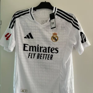 Vit Real Madrid fotbollströja Bellingham - Säljer en vit Real Madrid fotbollströja från Adidas i nytt skick. Tröjan har korta ärmar och är prydd med klubbens emblem och sponsorlogga. Baksidan har namnet 'Bellingham' och nummer 5. Storlek L