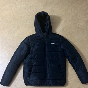 Svart pufferjacka från Jack & Jones - Säljer en svart pufferjacka från Jack & Jones i storlek M. Jackan är perfekt för kalla dagar med sin quiltade design och dragkedja framtill. Den har en praktisk huva och två fickor med dragkedja. Perfekt för både höst och vinter!