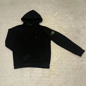 Stone Island hoodie - Säljer en snygg svart hoodie från Stone Island med deras ikoniska logga broderad på bröstet. Den har en klassisk känguruficka och en patch på ärmen. Perfekt för höst och vinter, både stilren och bekväm. Passar till alla tillfällen!