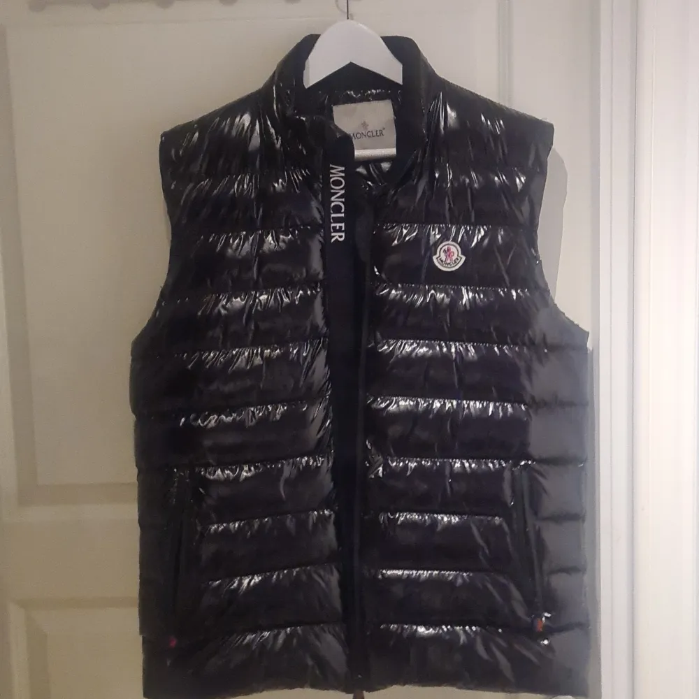 Säljer en snygg svart dunväst från Moncler i mycket bra skick. Aldrig använd. Västen har en glansig finish och är perfekt för höst och vinter. Den har en dragkedja framtill och Monclers ikoniska logga på bröstet. Insidan har en cool serietidningsdesign. Perfekt för att hålla sig varm och stilren!. Jackor.