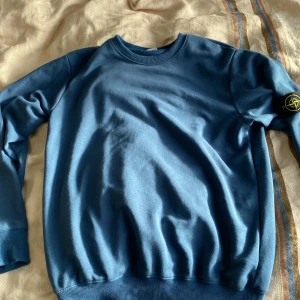 Blå tröja från Stone Island - Säljer en snygg blå tröja från Stone Island i storlek XL. Den har en klassisk rund hals och långa ärmar med det ikoniska märket på ärmen. Perfekt för både vardag och lite kyligare dagar. Tröjan är i mycket bra skick och redo för en ny ägare!