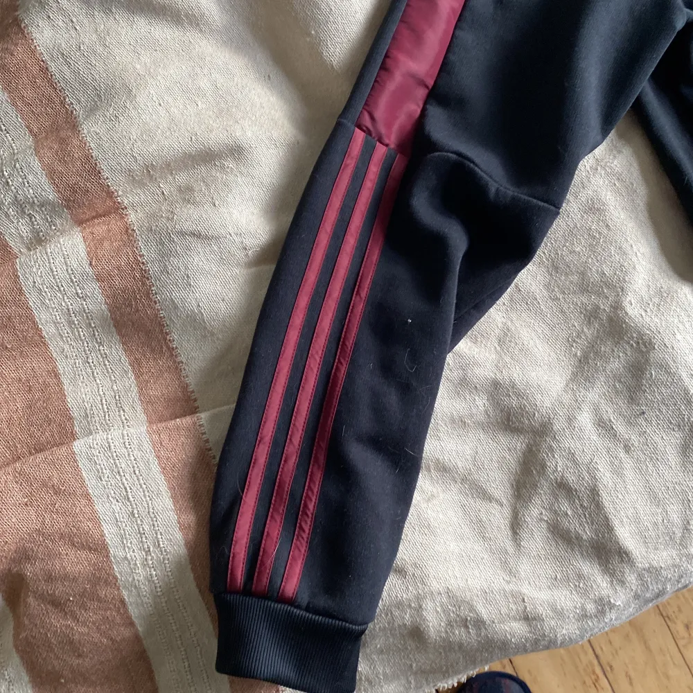 Säljer en snygg svart hoodie från Adidas med vinröda detaljer. Den har en halv dragkedja framtill och klassiska tre ränder längs ärmarna. Perfekt för höst och vinter, och den är i bra skick. Passar både för träning och vardagsbruk.. Tröjor & Koftor.