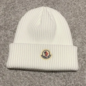 Vit ribbad mössa från Moncler - Säljer en snygg vit ribbad mössa från Moncler. Den är i nytt skick och perfekt för vintern. Mössan har en broderad Moncler-logga framtill och är supermjuk och bekväm. Passar perfekt till både vardag och skidresor! ❄️