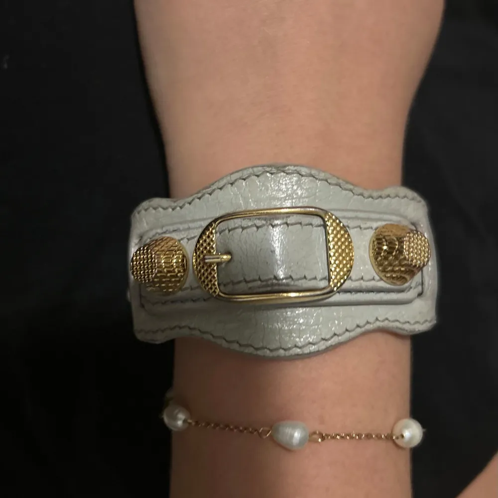 Snyggt grått armband från Balenciaga i skinn med guldiga detaljer. Armbandet har en unik design med ett spänne och texturen ger en lyxig känsla. Perfekt för att ge din outfit en exklusiv touch!. Accessoarer.