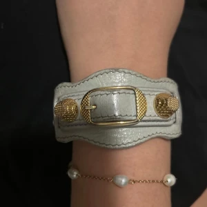 Grått armband från Balenciaga - Snyggt grått armband från Balenciaga i skinn med guldiga detaljer. Armbandet har en unik design med ett spänne och texturen ger en lyxig känsla. Perfekt för att ge din outfit en exklusiv touch!
