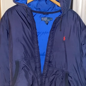 Blå vindjacka från Polo Ralph Lauren - Säljer en snygg blå vindjacka från Polo Ralph Lauren i storlek L. Jackan har en dragkedja framtill och är perfekt för höst och vår. Den är tillverkad i polyester och har en klassisk design med huva. Perfekt för dig som vill ha en stilren och funktionell jacka! 💙