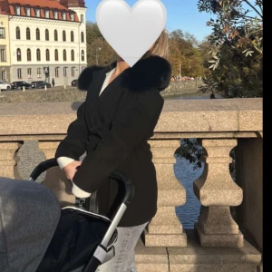 Svart kappa med pälsdetaljer - Säljer en elegant svart kappa med pälsdetaljer runt kragen. Perfekt för kyliga höst- och vinterdagar. Den har en klassisk passform och knyts i midjan för en snygg siluett. Passar både till vardags och festliga tillfällen.