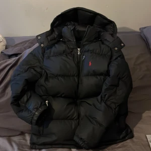 Svart jacka från Ralph Lauren - Säljer en svart dunjacka från Ralph Lauren i mycket bra skick. Jackan har en dragkedja framtill och en huva för extra värme. Perfekt för kalla vinterdagar! Den är långärmad och har en klassisk pufferstil med en liten röd logga på bröstet. Skick 10/10 alltså inga defekter. Storlek XS. Priset kan diskuteras😁👍