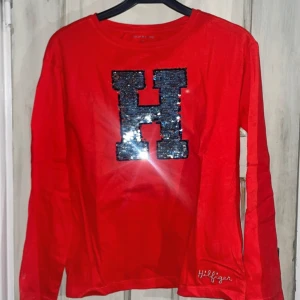 Röd långärmad tröja från Tommy Hilfiger - Säljer en snygg röd långärmad tröja från Tommy Hilfiger. Den har ett stort glittrigt 'H' på framsidan och Hilfiger-broderi nedtill. Tröjan är i fint skick och redo för en ny ägare! En ny tröja utan prislapp.