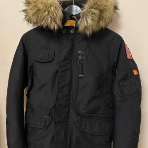 Svart junior vinterjacka från Parajumpers - Säljer en svart Parajumpers vinterjacka i perfekt skick. Jackan har en stor huva med pälsdetaljer och flera praktiska fickor. Perfekt för kalla vinterdagar.  Upphängaren i jackan har släppt på ena sidan (se bild) Storlek Young small ca 12-13år 152/158