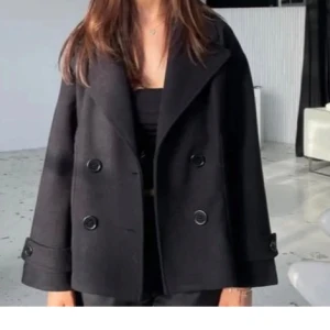 Svart kappa med knappar - Säljer en stilren svart kappa. Den har en klassisk design med stora knappar framtill och långa ärmar. Perfekt för höst och vår, och funkar både till vardags och fest. Passar till det mesta i garderoben! Negativa är att den har gått sönder i kappan men absolut ingen man ser utifrån 