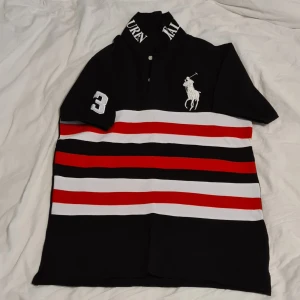 Sommar25. Mörkblå,röd,vit randig pikétröja från Ralph Lauren - Säljer en snygg pikétröja från Ralph Lauren i storlek L. Tröjan har ett klassiskt randigt mönster i svart, rött och vitt med en broderad logga på bröstet. Perfekt för en stilren och sportig look. Passar både till vardags och fest!
