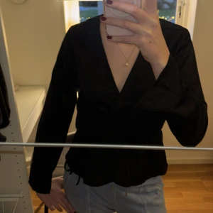 Svart omlottopp - Säljer en elegant svart omlottopp med långa ärmar. Toppen har en stilren design med knytning i midjan för en smickrande passform. Köptes på bikbok för ett år sedan 399kr. Aldrig använd och lappen finns kvar. 