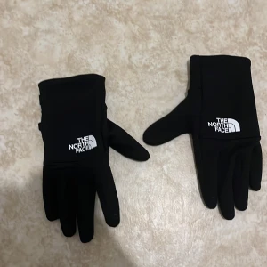 Svarta handskar från The North Face - Säljer ett par svarta handskar från The North Face i nytt skick. De är perfekta för höst och vinter, med en stilren design och logga på ovansidan. Handskarna har E-tip funktion för att använda mobilen utan att ta av dem. Perfekta för både vardag och utomhusaktiviteter!