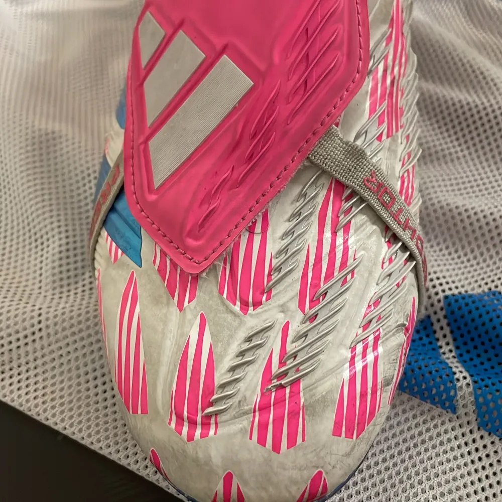 Säljer ett par Adidas Predator Elite fotbollsskor i vitt, rosa och blått. Skorna har en cool design med rosa och vita mönster och blå sula. De är i mycket bra skick då de inte används på mer än 3 veckor och ca 8 träningar och kommer med en praktisk förvaringspåse. Förvaringspåsen är lite sönder då ett av banden som håller upp skorna på ryggen är av. . Skor.