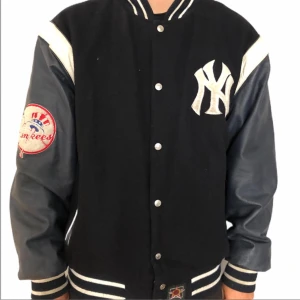 Svart New York bomberjacka Jeff Hamilton - Säljer en sällsynt vintage svart bomberjacka från Jeff Hamilton med New York Yankees-logga. Jackan har långa ärmar och knappar framtill. Den är gjord av polyester och läder, perfekt för höst och vår. Den är även reversible så man kan ha den på två sätt!                                                                    Yankees-märke på ärmen och text på ryggen ger en sportig look. Passar perfekt för casual outfits!  Perfekt för collectors!