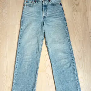 Säljer ett par Levi's Ribcage Straight jeans i ljusblå denim. De är högmidjade och har en klassisk femficksdesign med knappgylf. Perfekta för en avslappnad stil och passar till alla säsonger. Skicket är mycket bra, nästan som nya!