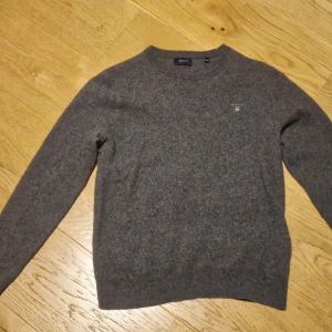 Grå ulltröja från GANT - Storlek L men i juniorstorlek så mer som XS för vuxen. Tröjan har en klassisk design med rund hals och GANT-logga på bröstet. Perfekt för både höst och vinter, och den är supermjuk och varm. 