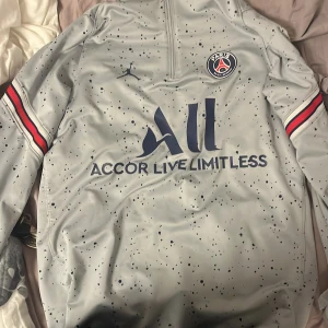 Grå PSG fotbollströja - Säljer en grå PSG fotbollströja från Jordan med svart och rött mönster. Tröjan har en halv dragkedja och klubbens emblem på bröstet. Perfekt för fans som vill visa sitt stöd! 🖤❤️