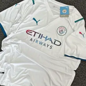 Säljer en vit Manchester City fotbollströja från Puma i nytt skick. Tröjan har korta ärmar med turkosa detaljer och klubbens emblem på bröstet. Perfekt för alla City-fans! 🩵