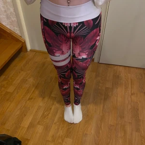 Rosa träningstights - Säljer ett par tights med ett blommönster i rosa och svart. Materialet är stretchigt och mjukt. De har också vita ränder på ena benet som påminner om tightsen från märket Stronger.
