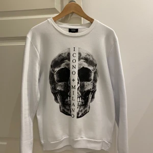 Icono Milano Sweatshirt - Säljer en stilren vit sweatshirt Tröja från Icono Milano med ett coolt dödskalletryck på framsidan. Tröjan har långa ärmar och en rund halsringning. Perfekt för en avslappnad och trendig look. Passar både till vardags och fest!