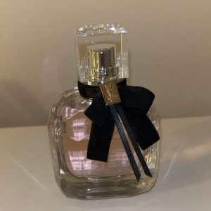 Parfym från Yves Saint Laurent - Säljer en helt ny bara utan förpackning elegant parfym från Yves Saint Laurent. Flaskan är genomskinlig med en lyxig svart rosett runt halsen och ett fyrkantigt lock. Perfekt för att ge en touch av glamour till din doftsamling.