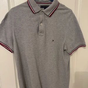 Grå pikétröja från Tommy Hilfiger - Säljer en stilren grå pikétröja från Tommy Hilfiger i storlek L. Tröjan har en klassisk krage med röd och vit randdetalj samt korta ärmar. Perfekt för både vardag och festliga tillfällen. Slim fit för en snygg passform!