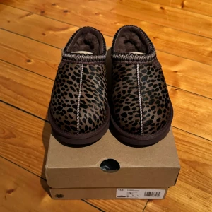 UGG tasman tofflor - Säljer ett par supermysiga leopardmönstrade tofflor från UGG. De är i brunt och svart med en mjuk insida som håller fötterna varma. Perfekta för att glida runt hemma under kalla dagar. De är helt nya och kommer i originalkartong. Finns i storlekarna 36, 38, 39, 40, 42.