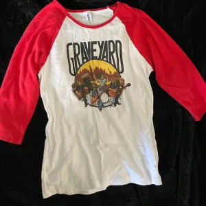 Långärmad band tshirt - Säljer en vit och röd t-shirt från Bella med ett Graveyard-tryck på framsidan. Bandet graveyard. Den har trekvartsärmar.