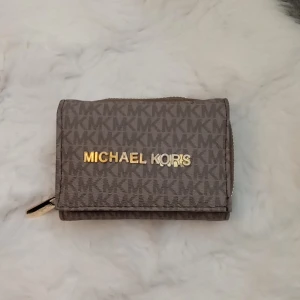 Brun plånbok från Michael Kors - Säljer en snygg brun plånbok från Michael Kors med det klassiska MK-mönstret. Plånboken har en praktisk dragkedja och flera fack för kort och kontanter. Perfekt för både vardag och fest! 👜✨