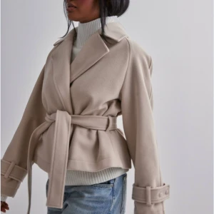 Beige kappa med bälte - OANVÄND MED PRISLAPP Beige kappa med bälte i midjan. Den har en  omlott-design och långa ärmar med detaljerade manschetter. Ganska stor i storleken så passar även s/m.  Ursprungs pris 699 kr och den är helt slutsåld på hemsidan. Pris kan diskuteras vid snabb affär