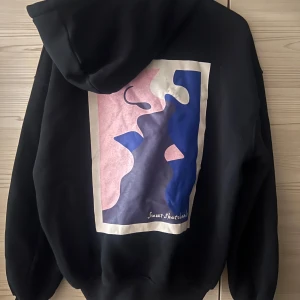 Svart hoodie med tryck - Säljer en svart hoodie med ett coolt konstnärligt tryck i rosa, blått och vitt på ryggen. Perfekt för höst och vinter, den är både stilren och bekväm. Hoodien har en klassisk passform med dragsko i huvan. Perfekt för att hålla sig varm och snygg! 🖤