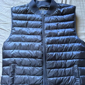 Svart dunväst från GANT - Säljer en snygg svart dunväst från GANT i storlek M. Västen har en dragkedja framtill och två praktiska fickor. Perfekt för höst och vår när du vill hålla dig varm men ändå se stilren ut. Passar både till vardags och lite finare tillfällen.