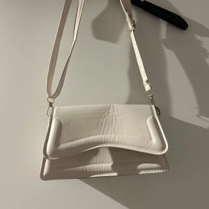 Vit beige axelväska i syntetmaterial - Säljer en stilren vit axelväska i syntetmaterial. Väskan har en justerbar axelrem och stängs med magnetknappar. Perfekt för både vardag och fest!