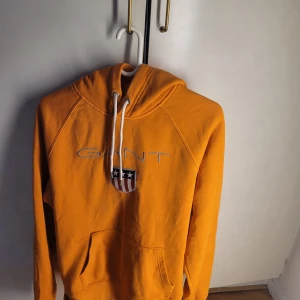 Orange hoodie från GANT - Säljer en snygg orange hoodie från GANT i bra skick. Den har en stor logga på bröstet och en praktisk magficka. Perfekt för höst och vår, med en skön passform och huva med dragsko. Passar både till vardags och chill.
