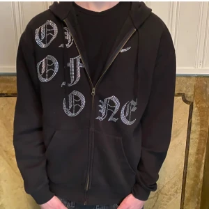 Svart hoodie från One of One - Säljer en svart hoodie från One of One i storlek medium. Den har en dragkedja framtill och är dekorerad med glittriga bokstäver. Perfekt för en cool och avslappnad stil. Riktigt fet och passar till dom flesta färgerna