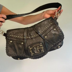 Svart axelväska med nitar och logotyp - Snygg svart vintage axelväska i skinn med coola nitar och en stor logotyp på framsidan. Väskan har en praktisk dragkedja på baksidan och ett mönstrat innerfoder. Perfekt för både vardag och fest!           Bilderna visar lite tydligare vilket skick väskan är i!