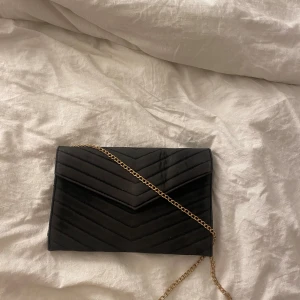 Svart quiltad kuvertväska med kedja - Snygg svart kuvertväska i quiltat mönster med en elegant guldkedja som axelrem. Perfekt för en utekväll eller fest! Väskan har en praktisk magnetknapp för enkel öppning och stängning. Stilren och chic design som passar till alla outfits.