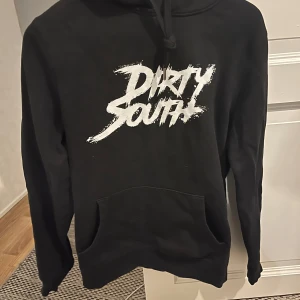 Svart hoodie med tryck - Säljer en svart hoodie med texten 'Dirty South' i vitt på bröstet. Den är i bra skick och perfekt för höst och vinter. Hoodien har en klassisk passform med långa ärmar och en stor ficka framtill. Perfekt för en avslappnad stil! Är gjord av rapparen dizzy