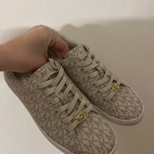 Beiga Michael Kors skor med monogram. - Säljer ett par snygga Michael Kors skor med monogrammönster. De har snörning och en liten gulddetalj framtill. Perfekta för en stilren vardagslook! Jag har endast använt dessa skor upp till 2-4 gånger, de är i mycket bra skick och kan ses som nya.