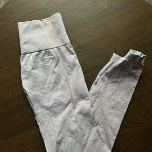 Ljuslila leggings med ribbad midja - Säljer ett par ljuslila leggings i mycket bra skick. De har en ribbad hög midja och är perfekta för träning. Materialet är mjukt och stretchigt, vilket gör dem superbekväma. Det finns en diskret logga på benet. Perfekta för gymmet eller yoga!