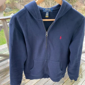 Mörkblå hoodie från Ralph Lauren - Säljer en snygg mörkblå hoodie från Ralph Lauren i storlek 14-16/Xs. Den har en dragkedja framtill och den klassiska röda loggan på bröstet. Perfekt som julklapp. Grymt skön och stilren!