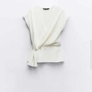 Vit omlottopp från Zara - Snygg vit omlottopp från Zara i ett mjukt och bekvämt material. Toppen har en elegant knytning i sidan och är ärmlös, vilket gör den perfekt för både vardag och fest. Passar bra till både jeans och kjol!