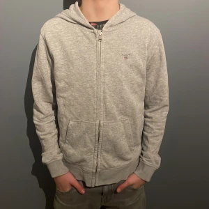 Grå hoodie från GANT - Säljer en snygg och bekväm grå hoodie från GANT. Den har en dragkedja framtill och en broderad logga på bröstet. Perfekt för både vardag och chill. Den är i bra skick och passar till det mesta. Hör av dig om du är intresserad!