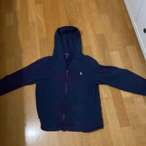 Mörkblå hoodie från Ralph Lauren - Säljer en snygg mörkblå hoodie från Ralph Lauren i storlek XL barn som är som storlek xs-s Den har en dragkedja framtill och den klassiska loggan broderad på bröstet. Perfekt för höst och vinter, och den är i mycket bra skick. Kan gå ner i pris vid snabb affär 