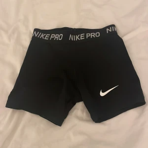 Svarta träningsshorts från Nike - Säljer ett par svarta Nike Pro träningsshorts i mycket bra skick. De är tighta och perfekta för träning. Shortsen har en elastisk midja med Nike Pro-logga och en vit Nike-swoosh på benet. Superbekväma och stilrena!