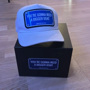 Vit keps från John Hatter & Co - Snygg vit keps med ett blått märke där det står 'You're gonna need a bigger boat'. Perfekt för sommarens äventyr eller bara för att se cool ut. Kepsen har en klassisk passform och är tillverkad i bomull med mesh-detaljer för extra ventilation.