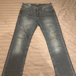 Blå jeans från Jack & Jones - Säljer ett par snygga blå jeans från Jack & Jones i storlek 30/32. De har en klassisk straight fit och är i bra skick med några coola slitningar på framsidan. Perfekta för en avslappnad stil till vardags eller fest. 🕺